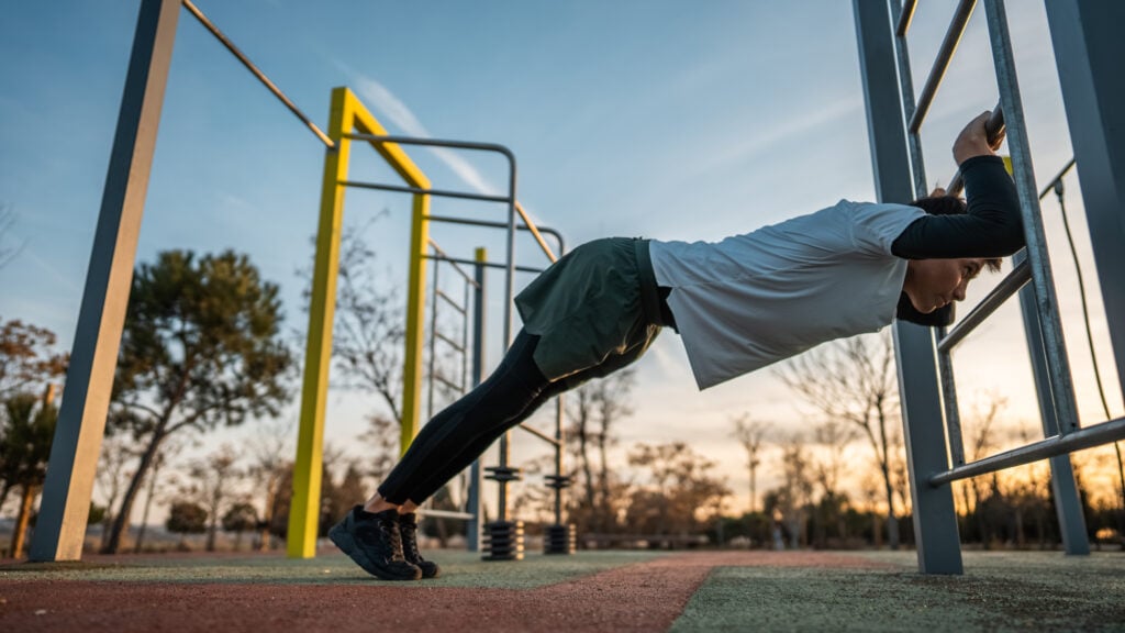 young athletic man exercising at fitness calisthenics workout and stretching legs and back at street gym during sunset, body improvement and strength training concept